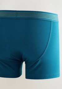 Türkisblaue Herren Boxershorts aus dehnbarem Stoff, mit glatter Textur, kurzen Beinen und einem Bund mit goldenen Streifen.