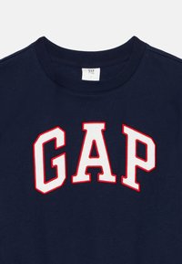 Tengerészkék pamut pulóver, amelyen egy merész fehér "GAP" logó látható, piros kerettel. Kerek nyakú dizájn, puha, texturált felülettel.