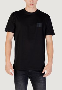 Camiseta negra de manga corta hecha de algodón, con un pequeño parche texturizado en el lado izquierdo del pecho y un diseño de cuello redondo clásico.