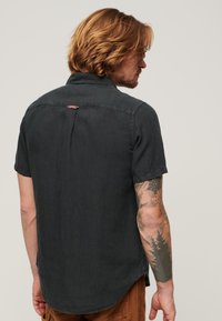 Camicetta nera a maniche corte realizzata in tessuto leggero. Presenta un colletto classico, una piega sulla schiena e una targhetta con il marchio vicino al colletto.