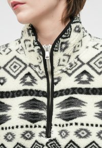 Pūkų megztinis su juodai baltu geometriniu raštu, aukštu apykakle ir sidabriniu priekinio užtrauktuko užsegimu. Minkšta tekstūra su kontrastinėmis juodomis detalėmis.