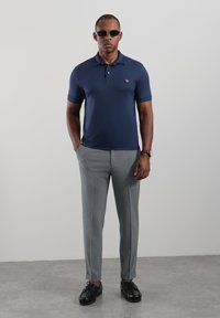 Granatowa koszulka polo z logo, jasnoszare spodnie na miarę oraz czarne buty. Strój ma dopasowany krój i gładką fakturę.