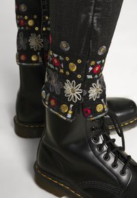 Botas negras de cuero con cordones combinadas con pantalones negros que presentan coloridos bordados de flores y lentejuelas doradas alrededor de los puños.