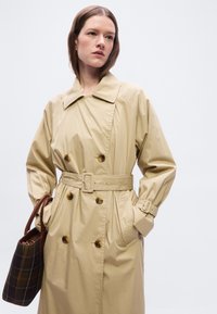 Femme portant un trench-coat beige ceinturé avec des boutons en écaille de tortue, tenant un sac fourre-tout à carreaux marron, debout devant un fond uni.