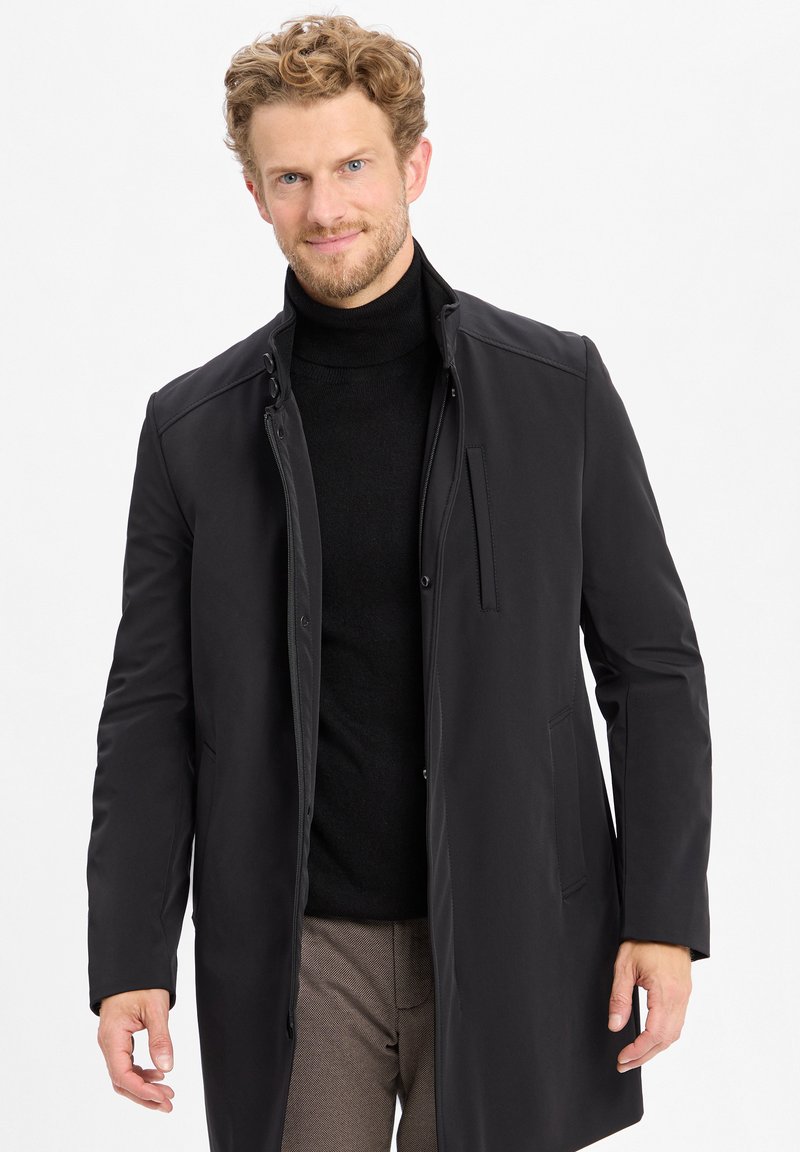 Zwarte lichtgewicht jas met drukknoopsluiting en hoge kraag. Extra ritssluiting detail op de voorkant, gedragen over een zwart coltrui.