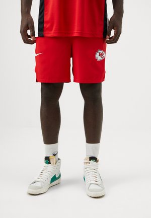 Homme portant un short rouge des Kansas City Chiefs, des baskets blanches Nike avec un swoosh vert et des chaussettes blanches sur un fond uni.