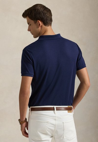Mann, der ein dunkelblaues Poloshirt mit kurzen Ärmeln, weiße Hose und einen braunen Ledergürtel trägt, von hinten vor einem schlichten Hintergrund gesehen.