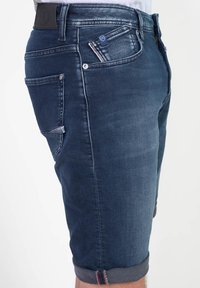 Shorts de mezclilla azul oscuro con corte ajustado, dobladillos enrollados y cinco bolsillos. Presentan un parche de cuero en la cinturilla y un ligero desgaste.