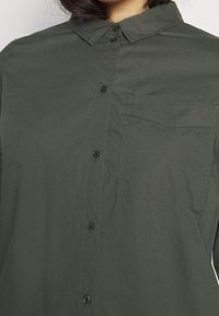 Dark green button-up shirt made of smooth fabric, featuring a collar, front pocket, and black buttons along the placket.