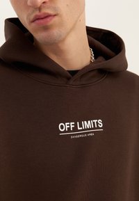 Felpa marrone con cappuccio, con stampa frontale che recita "OFF LIMITS" e "DANGEROUS AREA" in bianco. Texture morbida, design casual e cappuccio con coulisse.