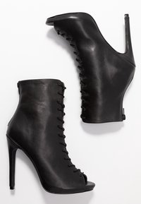 Bottines à talons hauts noires en cuir avec des lacets à l'avant et bout découvert, présentées sur un fond blanc.