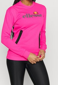 Felpa rosa con tasche zip nere, polsini e fondo a costine, e grafica del logo sul davanti. Texture liscia, vestibilità atletica.