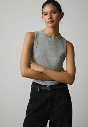 Young woman with arms crossed wearing a sleeveless gray top and black pants with a belt, standing against a plain gray background.