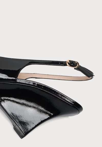 Μαύρο παπούτσι slingback από δέρμα με γυαλιστερή επίστρωση, μυτερή μύτη, μικρό σφήνα τακουνιού και χρυσή πόρπη στο λουράκι του αστραγάλου, σε λευκό φόντο.
