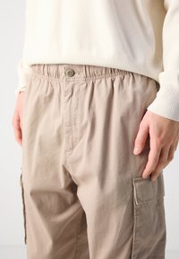 Pantaloni cargo bej cu talie elastică, închidere cu nasture și detalii cu buzunare laterale. Materialul pare ușor și ușor șifonat.