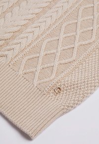 Tissu tricoté couleur crème avec motifs torsadés et diamants, bord côtelé, et petit logo de marque métallique circulaire près du coin inférieur droit.