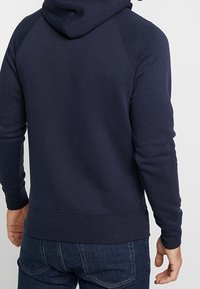 Uomo che indossa una felpa con cappuccio blu navy e jeans blu scuri, visto da dietro su uno sfondo chiaro e uniformi.