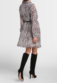 Woman wearing a knee-length, long-sleeve patterned dress and black high-heeled knee boots, standing against a light background.