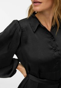 Melnā blūze ar gludu, spīdīgu tekstūru. Tai ir pogu aizdare priekšpusē, smaili izgriezts apkakle un apjomīgas piedurknes. Komplektā ietilpst atbilstoša josta.