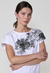 Camiseta blanca de manga corta con un estampado abstracto negro en el hombro izquierdo, hecha de suave tela de algodón. Textura lisa y cuello redondo.