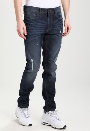 Homme portant un jean skinny bleu foncé usé et des baskets blanches de sport, debout sur un fond clair.