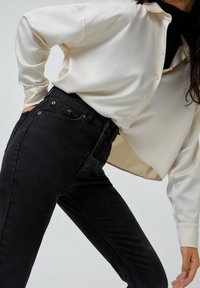 Camisa abotonada de satén blanco con mangas largas, combinada con unos jeans de mezclilla negros de cintura alta que cuentan con herrajes de cobre y una silueta ajustada.