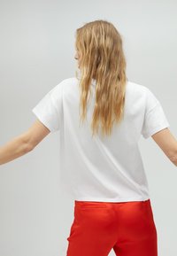 Bílá košile s krátkým rukávem a uvolněným střihem, spárovaná s červenými kalhotami. Košile má jednoduchý design a měkkou texturu.