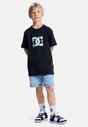 Czarny bawełniany t-shirt z zielono-białym logo DC, zestawiony z jasnoniebieskimi szortami jeansowymi, białymi skarpetkami oraz czarno-czerwonymi sneakersami.