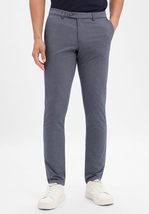 Mężczyzna w niebieskich, dopasowanych spodniach z przodu na guziki i zamek, noszący białe trampki i trzymający ręce w kieszeniach.