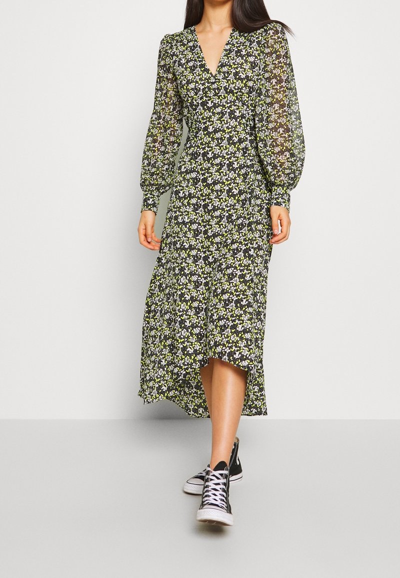 Femme portant une robe fleurie à manches longues avec un décolleté en V et des baskets montantes noires, debout devant un fond clair uni.