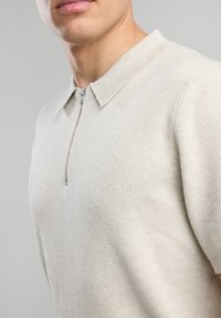 Vaaleanbeige lyhythihainen polo-paita, jossa on ribbikuvio ja vetoketjukaulus. Kangas näyttää pehmeältä ja istuvalta, korostaen esteettisesti siistiä muotoilua.