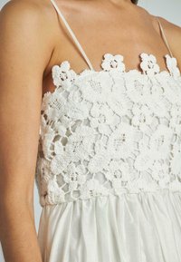 Witte jurk met een gehaakt bloemenpatroon aan de bovenkant en een gladde, glanzende stof in de rok. Spaghetti-bandjes maken de look af.