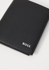 Portefeuille en cuir noir avec une surface texturée. Logo "BOSS" en argent à l'avant. Conception pliée avec des bords nets.