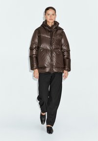 Hnědá péřová bunda s kapucí, lesklý materiál, prošívaný design, přední zip a boční kapsy. Vytvořeno s černými kalhotami a botami.