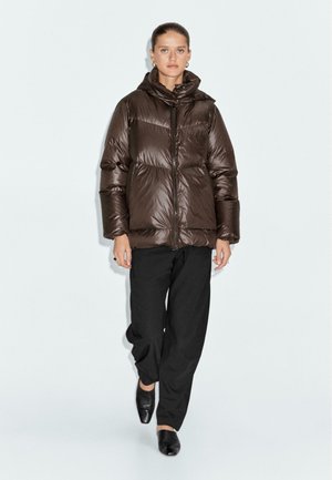 Giacca piumino marrone con cappuccio, materiale lucido, design trapuntato, zip frontale e tasche laterali. Abbinata a pantaloni neri e scarpe.