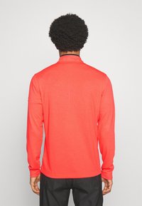 Un homme aux cheveux bouclés portant une chemise rouge vif à manches longues et un pantalon noir, se tenant dos à l'appareil photo devant un fond blanc.
