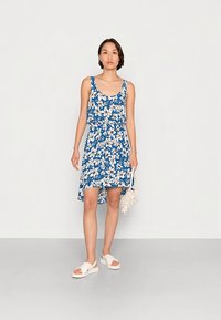 Mulher vestindo um vestido azul sem mangas com estampa floral e barra assimétrica, segurando uma mala branca texturada e usando sandálias brancas, em pé contra um fundo simples.