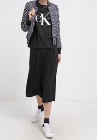 Vrouw in zwarte Calvin Klein T-shirt, met een jas van luipaardprint, wijde zwarte culottes, zwarte sokken en witte sneakers die vooruit loopt.