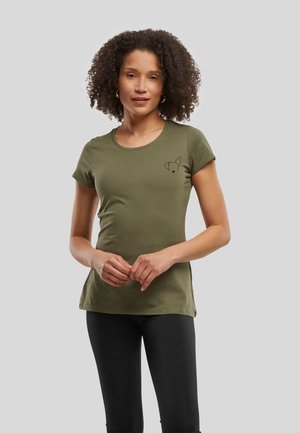 Femme aux cheveux bouclés porte un t-shirt vert olive avec un petit motif de visage de chien et un pantalon noir, debout devant un fond gris.