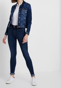 Tmavě modrá džínová bunda s předními kapsami a zapínáním na knoflíky. Kombinována s těsně padnoucími tmavými džínami a bílými teniskami.
