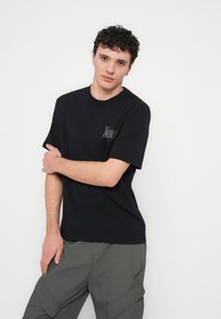 Črna bombažna majica s kratkimi rokavi, z diskretnim logotipom na prsih. Tkanina je gladka, z udobnim, sproščenim krojem.