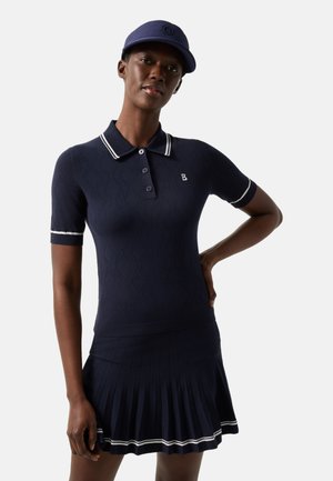 Vrouw draagt een marineblauw poloshirt met korte mouwen en een geplooide rok met witte rand, gecombineerd met een bijpassende marineblauwe honkbalpet.