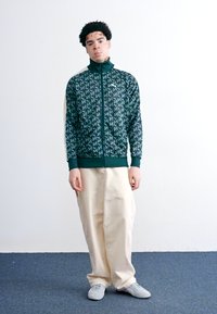 Veste zippée verte avec un motif logo intégral, associée à un pantalon beige ample et des baskets grises. Fond blanc uni.