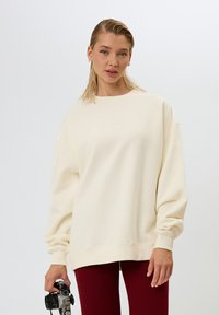 Felpa oversize color crema realizzata in morbido tessuto, con scollo rotondo e polsini a costine, abbinata a legging bordeaux a costine e una fotocamera.