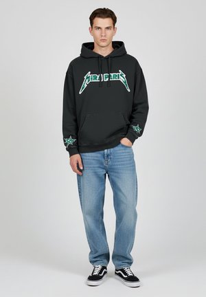 Zwarte hoodie met groene grafische logo en steraccenten, gecombineerd met lichtblauwe spijkerbroek en zwarte sneakers met witte zolen.
