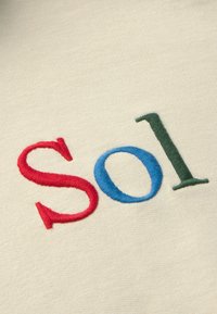 Embroidered letters "S" in red, "o" in blue, and "I" in green on a beige fabric background, showcasing a soft texture and clean design.