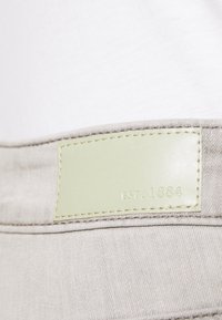 Una tela gris con una textura suave presenta un parche de cuero verde claro etiquetado como "EST. 1884" cosido en la cinturilla.