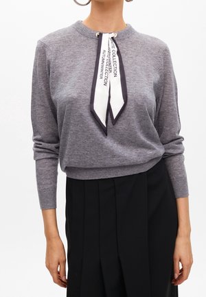 Vrouw met een grijze trui met lange mouwen en een zwart-witte sjaal geknoopt om de hals, gecombineerd met een zwarte plissérok.