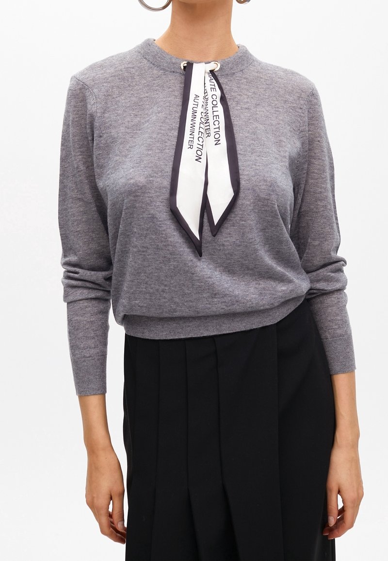 Femme portant un pull gris à manches longues avec une écharpe noire et blanche nouée autour du cou, assorti d'une jupe plissée noire.