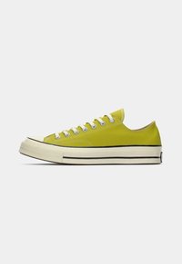 Adidaș galbeni din pânză cu șireturi albe, vârf de cauciuc și o dublă bandă neagră pe talpă. Design cu talpa joasă, textură netedă și formă rotundă.